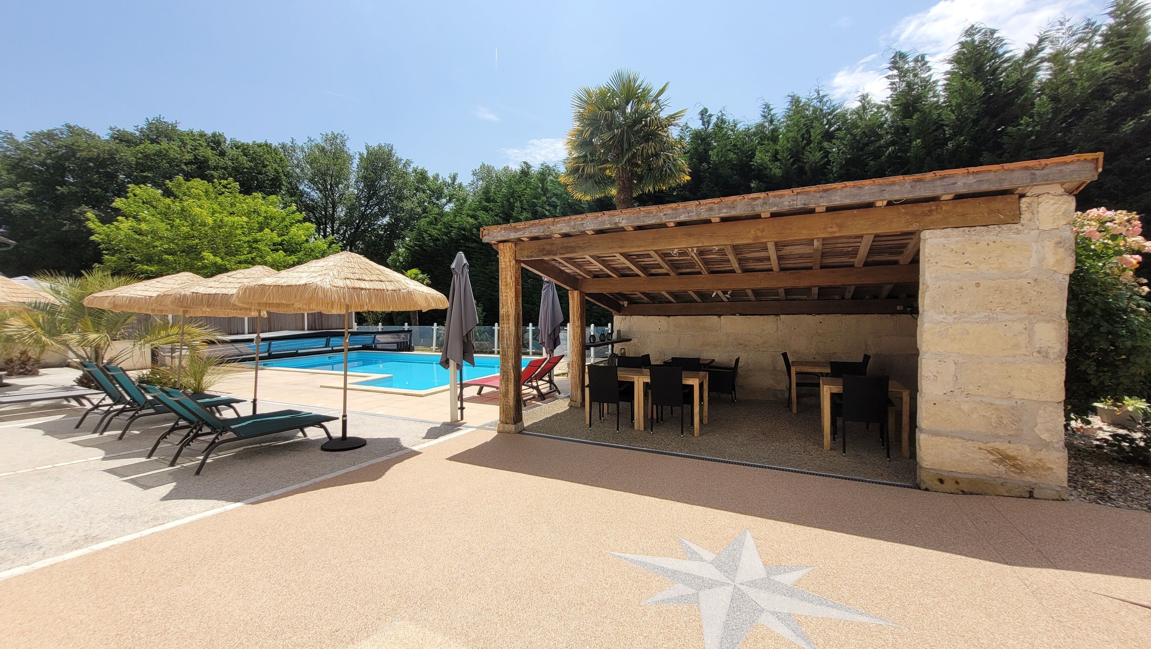A pool area with lounge chairs and umbrellas, a covered seating area with tables, and a stone wall.