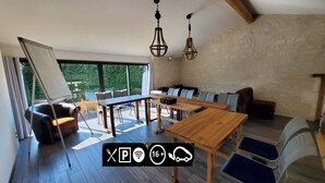 A spacious room with a wooden table, chairs, and a sofa. There are two hanging lights and a large window providing a view of greenery outside. - Les Cuves (Chaniers)