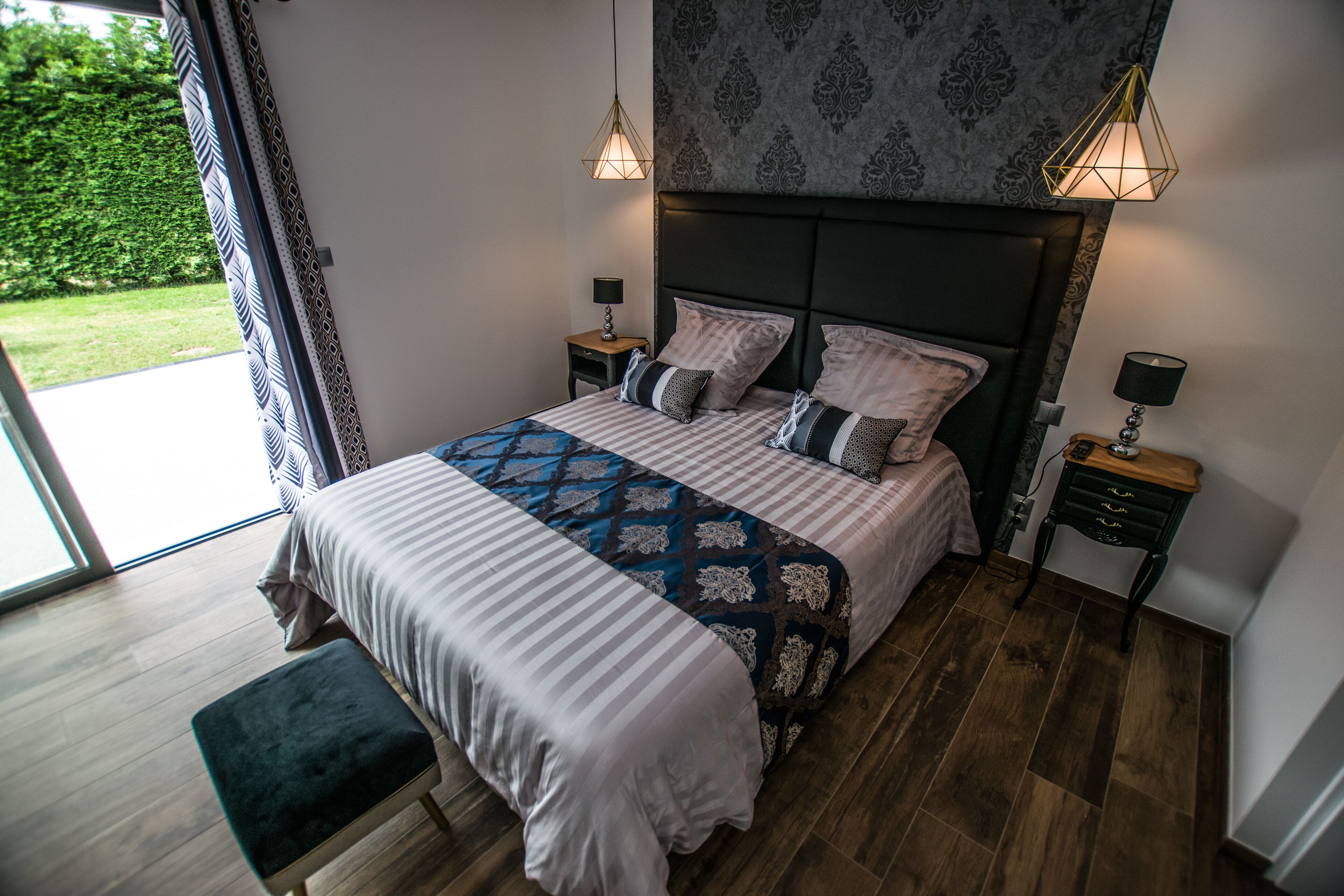 A bedroom with a large bed, two bedside tables with lamps, a bench, and a view of a garden through a sliding door.
