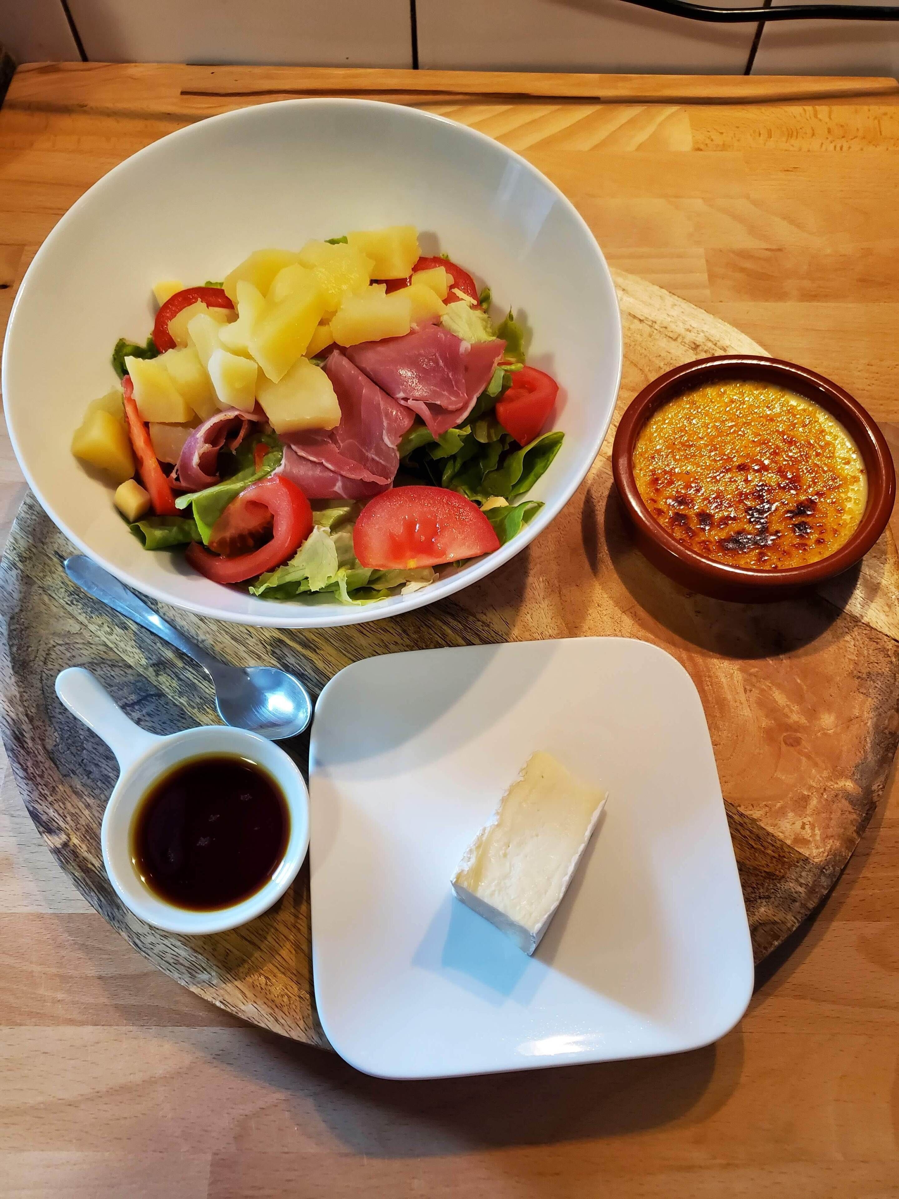 Ein Holztisch mit einer weißen Schüssel, gefüllt mit Salat, einer kleinen Schale mit Sauce und einem Käsewürfel auf einem weißen Teller.