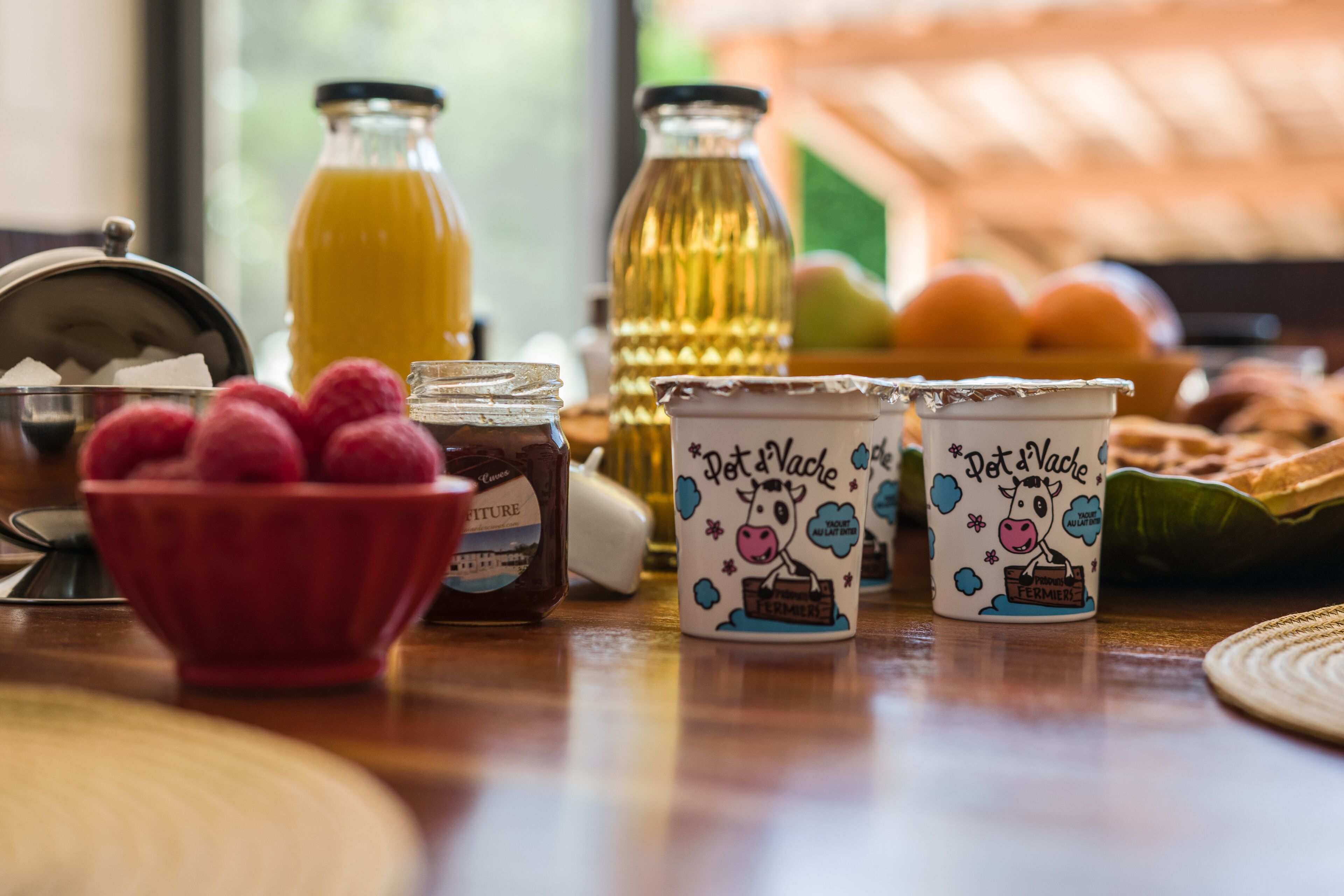 Ein Frühstückstisch mit einer Schüssel Himbeeren, einem Marmeladenglas, einer Orangensaftflasche und zwei Tassen mit einem Bauernhoftier-Motiv.