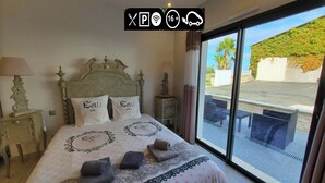 A hotel room with a large bed, two pillows with "Lau de" text, a nightstand with a lamp, and a view of the outside through a large window. - Les Cuves (Chaniers)