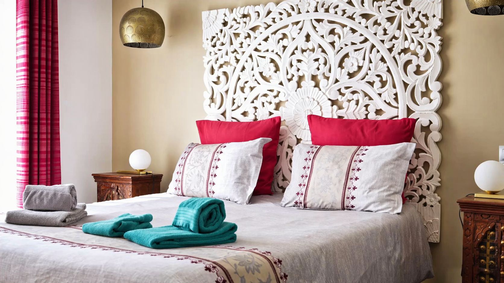 A neatly made bed with red and white pillows, a decorative headboard, and a pair of towels on the floor.