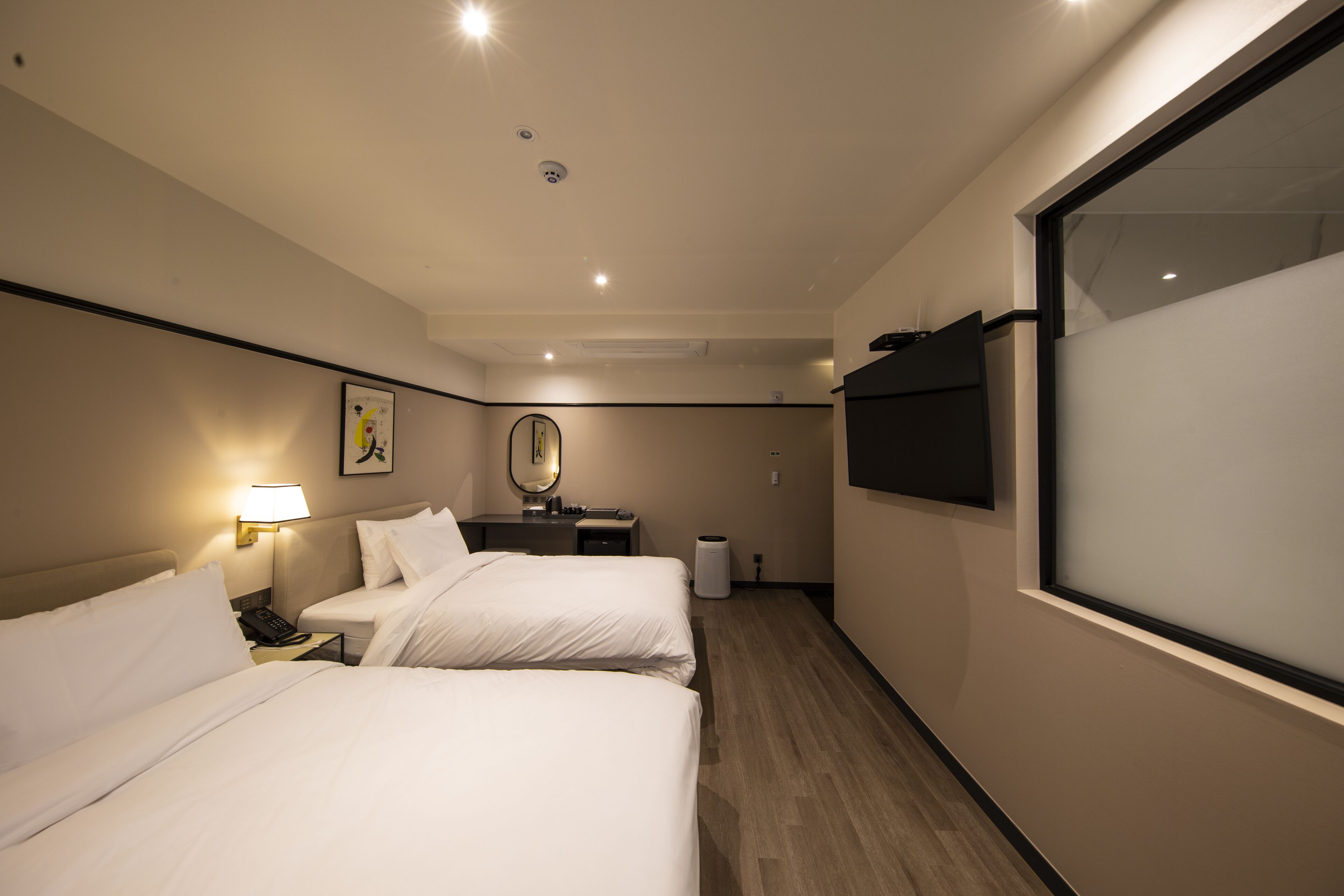 A hotel room with two beds, a bedside table with a lamp, a mirror, a TV mounted on the wall, and a waste bin.