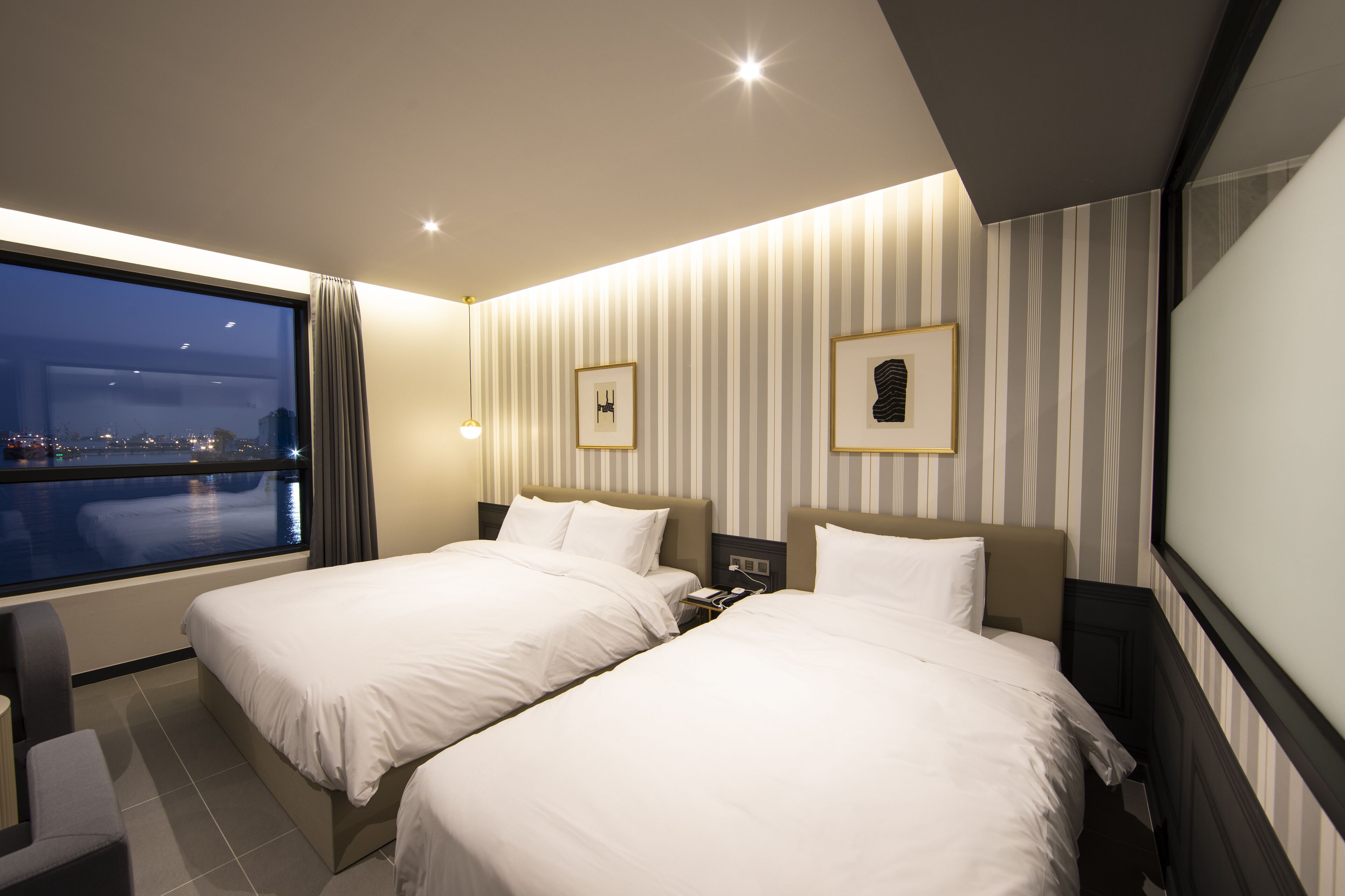 A hotel room with two beds, a large window with a city view, striped wallpaper, and a night sky visible through the window.