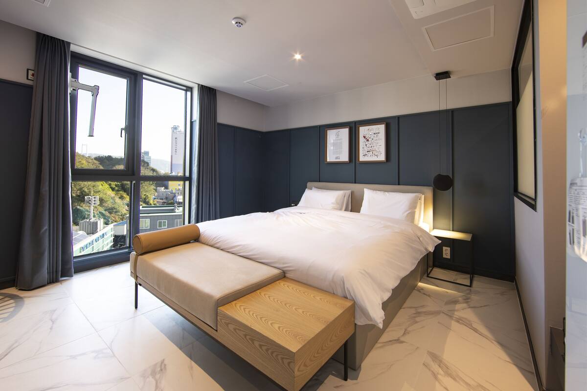 Ein modernes Schlafzimmer mit einem großen Bett, einer Bank und Blick auf Gebäude durch ein großes Fenster.