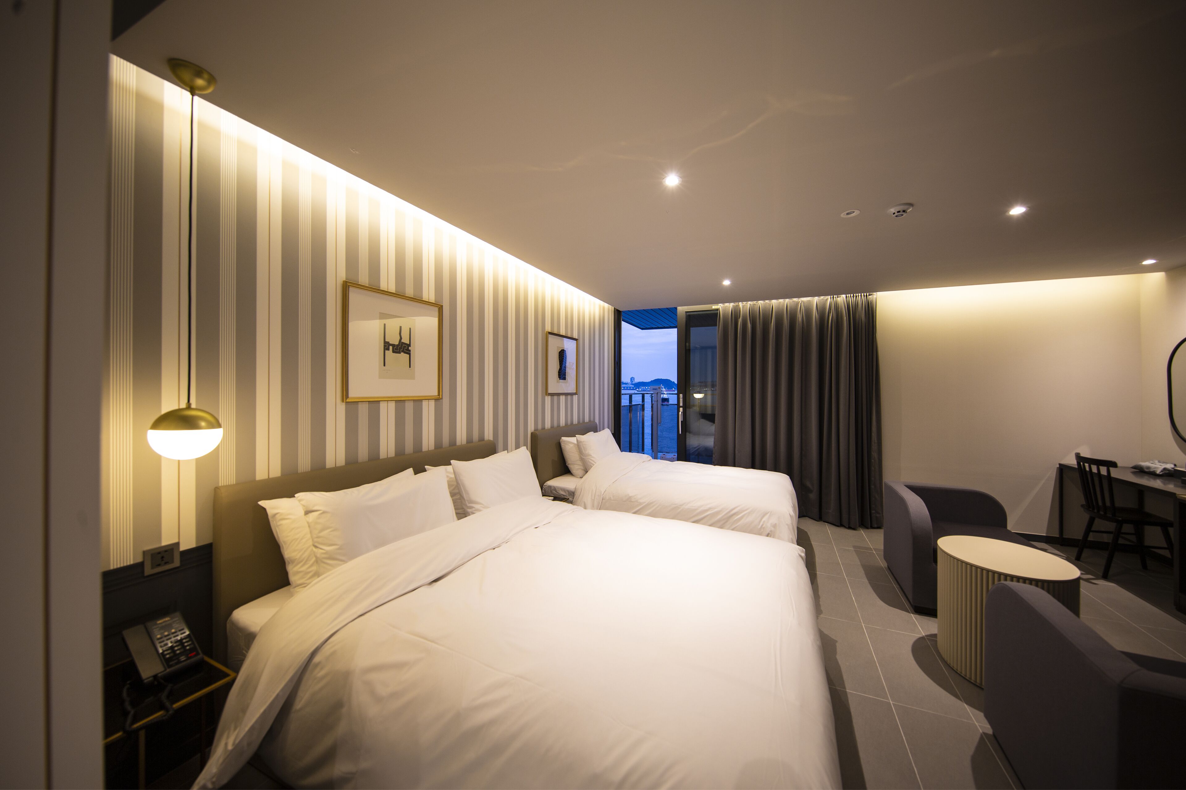 A hotel room with two beds, a desk, a chair, a lamp, and a view of the city.