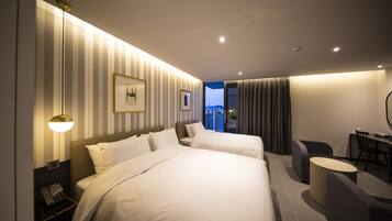 A hotel room with two beds, a desk, a chair, a lamp, and a view of the city.