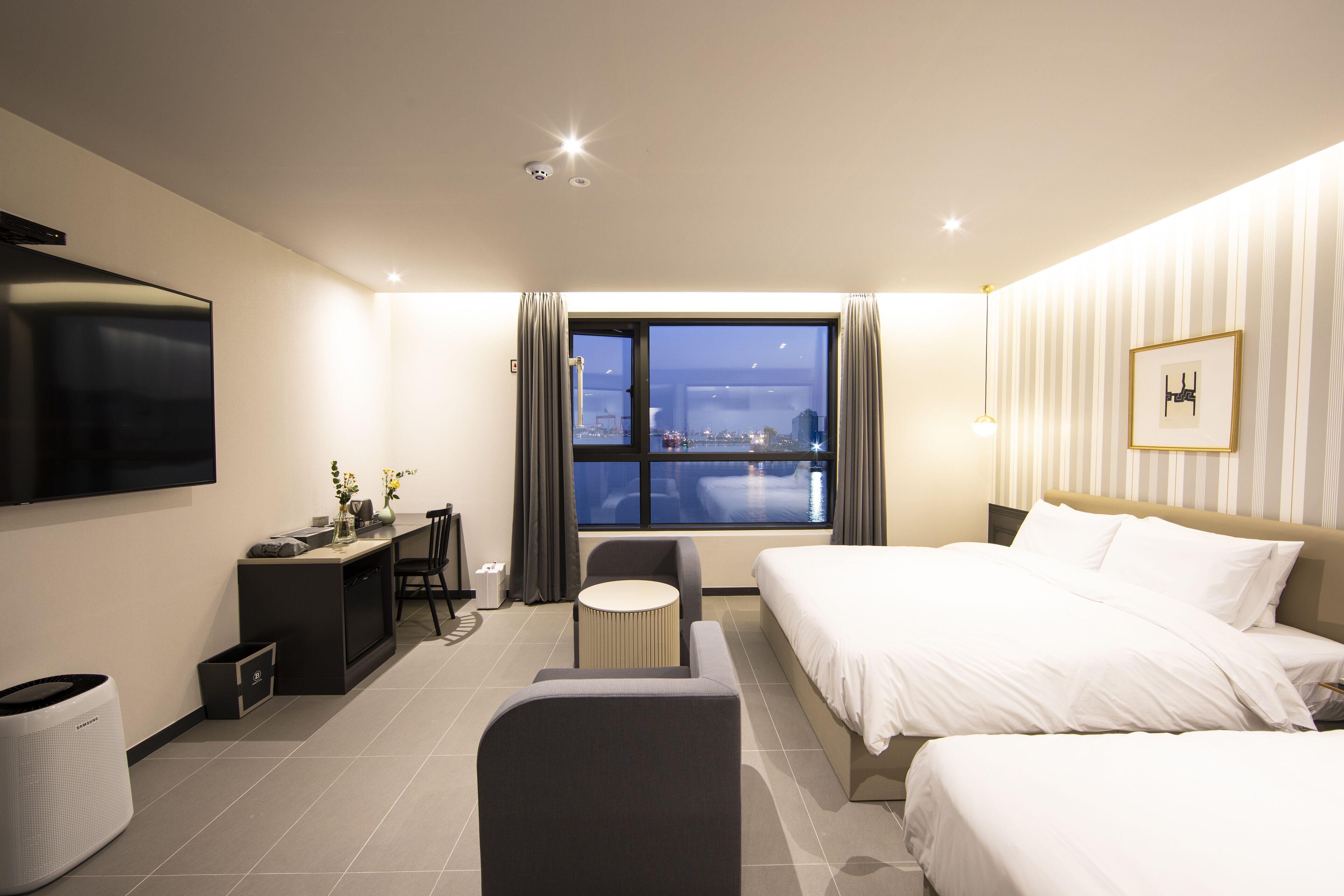 A modern hotel room with a large bed, a desk with a chair, a small round table, a TV mounted on the wall, and a window with a city view.