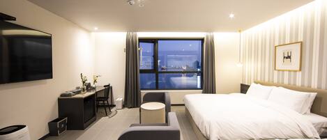 A modern hotel room with a large bed, a desk with a chair, a small round table, a TV mounted on the wall, and a window with a city view.