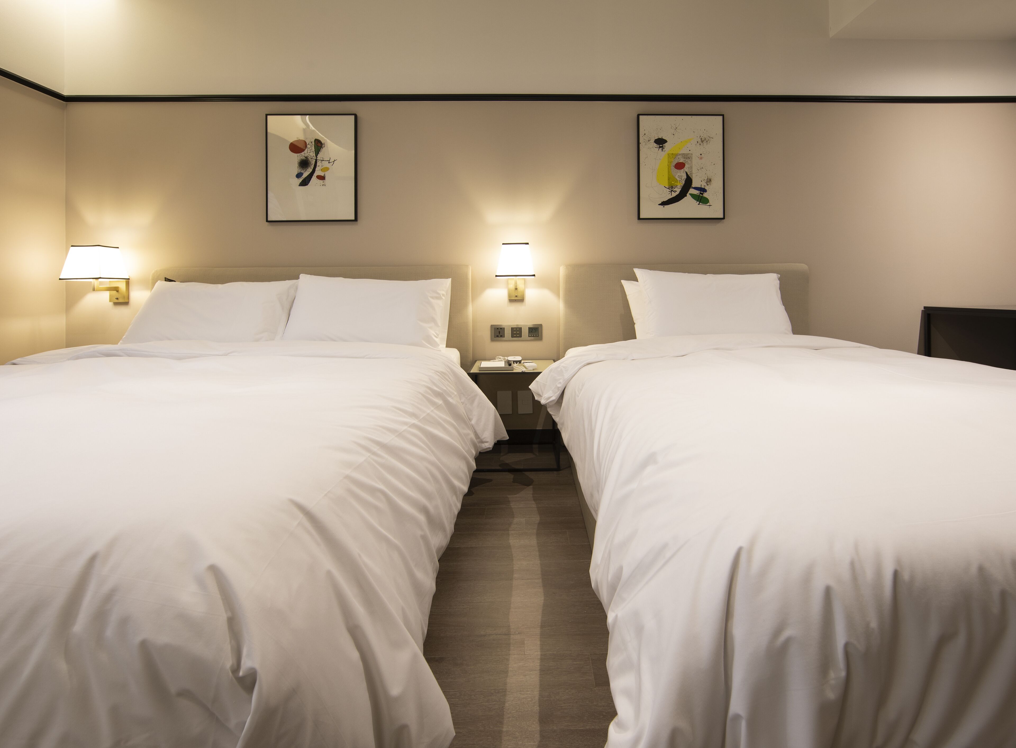 Habitación de hotel con dos camas, cada una con ropa de cama blanca, dos lámparas fijadas a la pared y dos cuadros abstractos enmarcados.