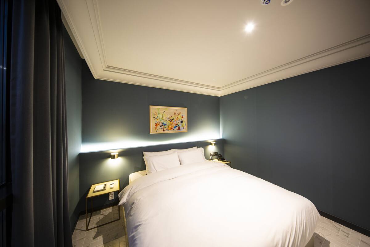 Ein ordentlich bezogenes Bett in einem Zimmer mit dunklen Wänden, ein Bild an der Wand und Nachttischlampen.