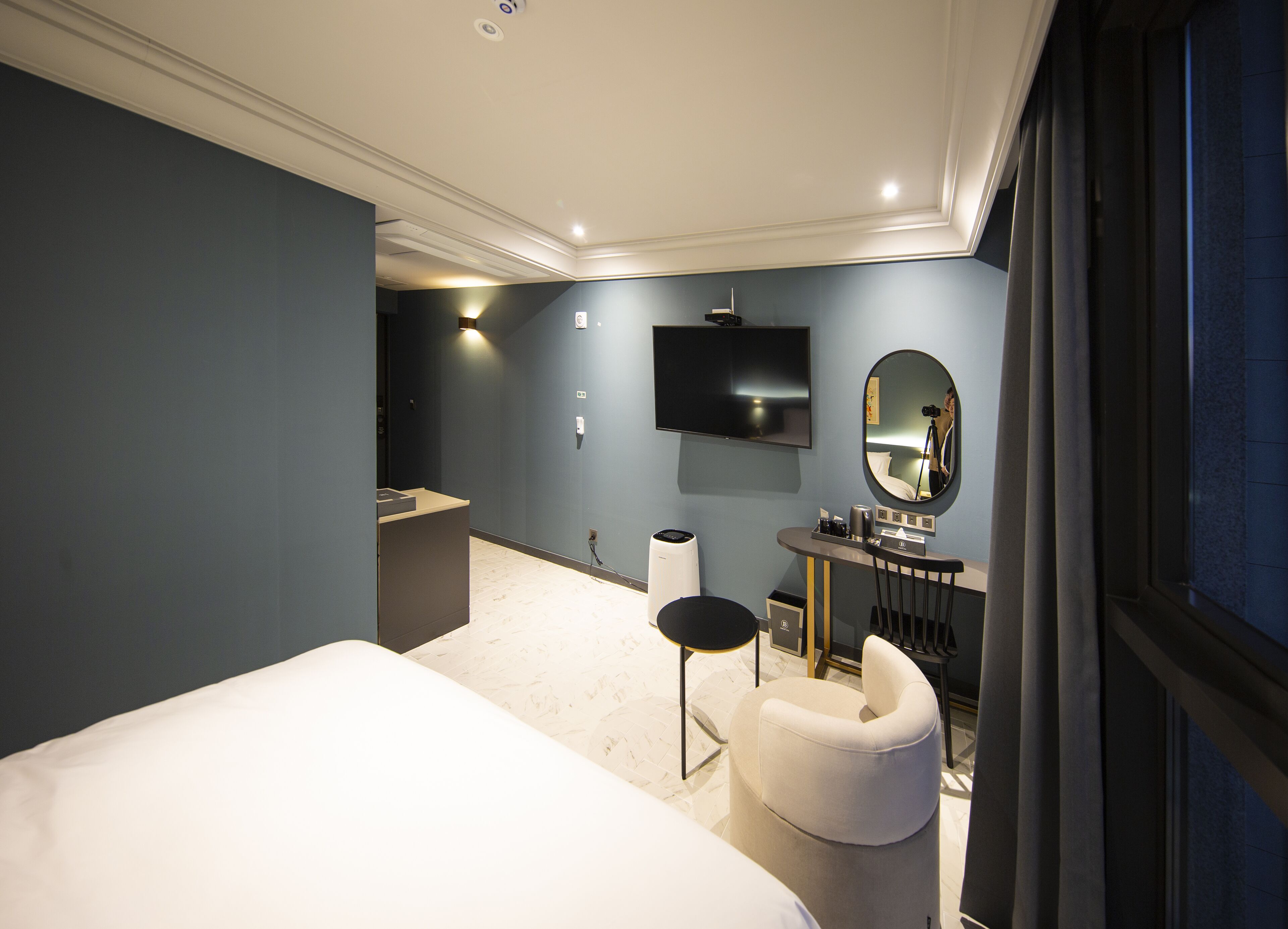 A modern hotel room with a large bed, a vanity with a mirror, a flat-screen TV, and a seating area with a chair and a small table.