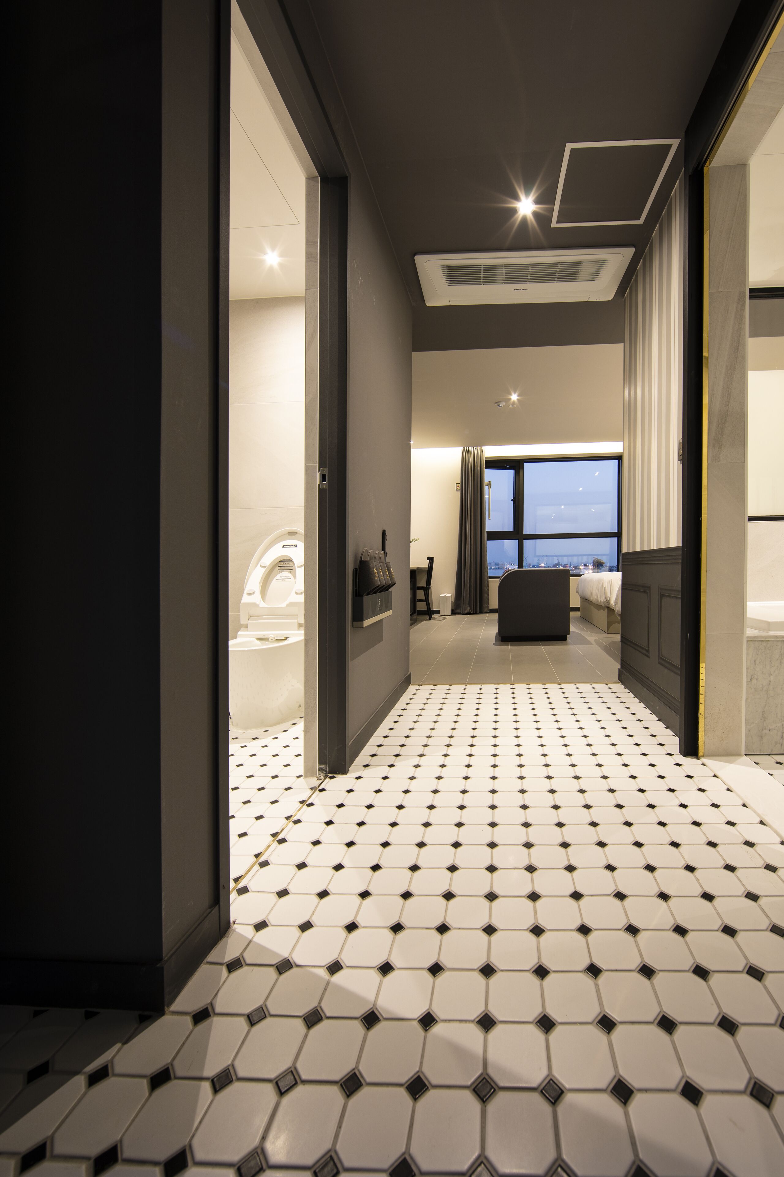 A modern hotel room with a geometric tile floor, a large window with a city view, a bathtub, and a bed.