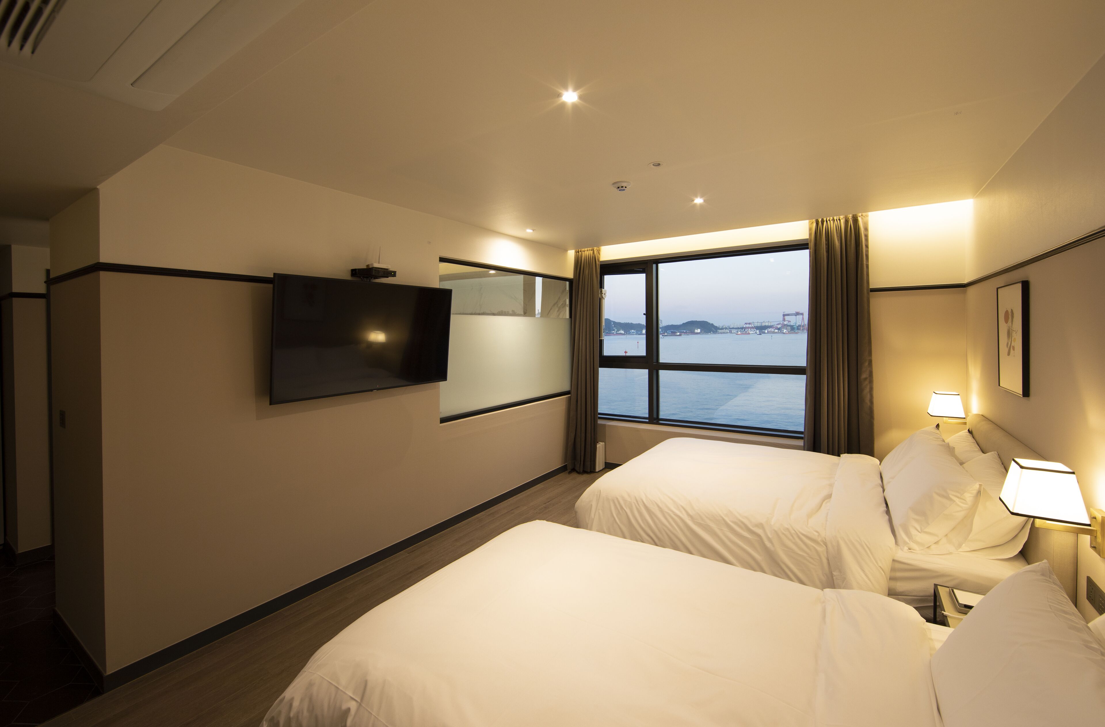 Une chambre d’hôtel avec deux lits, un téléviseur fixé au mur, une fenêtre donnant sur la mer et une table de chevet avec une lampe.