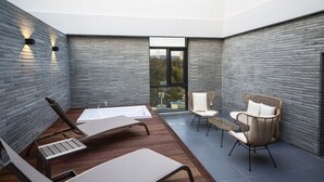 Een modern terras buiten, met een houten tafel en stoelen, een rieten fauteuil en uitzicht op een brug door een glazen deur.