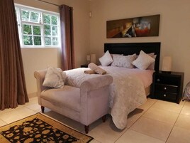 Quarto com cama, duas poltronas, mesa de cabeceira, pintura e janela com cortinas.
