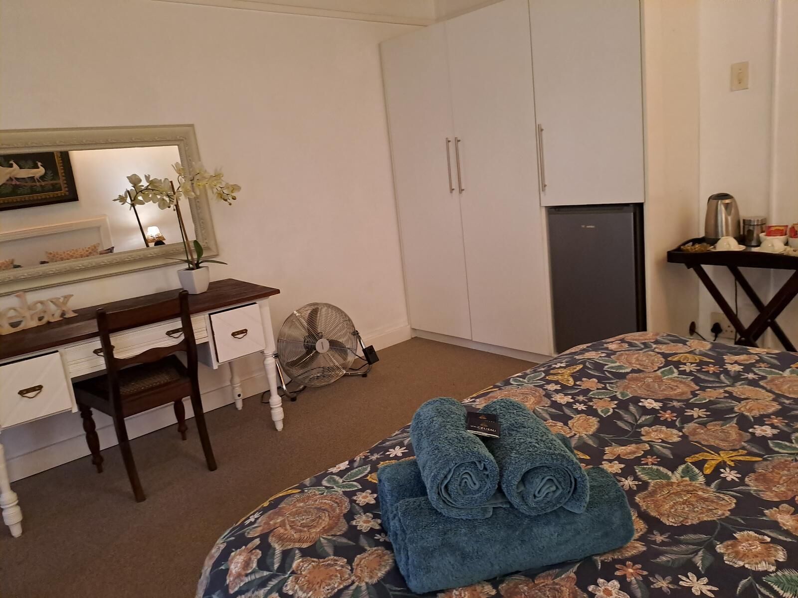 Habitación de hotel con una cama, un escritorio con espejo, un aparador, un ventilador y un refrigerador pequeño.