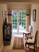 Um corredor com tapete estampado, duas cadeiras, uma mesa pequena e um armário com objetos decorativos.