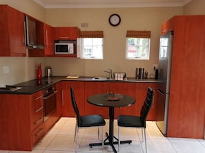 A compact kitchen with wooden cabinets, a microwave, a refrigerator, a dining table with chairs, and a clock on the wall.