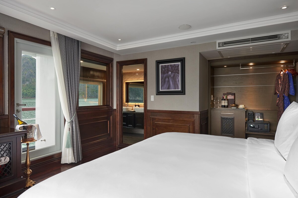 Habitación de hotel con una cama grande, un minibar, un escritorio con lámpara y vistas al mar. Se ve un baño por una puerta abierta.