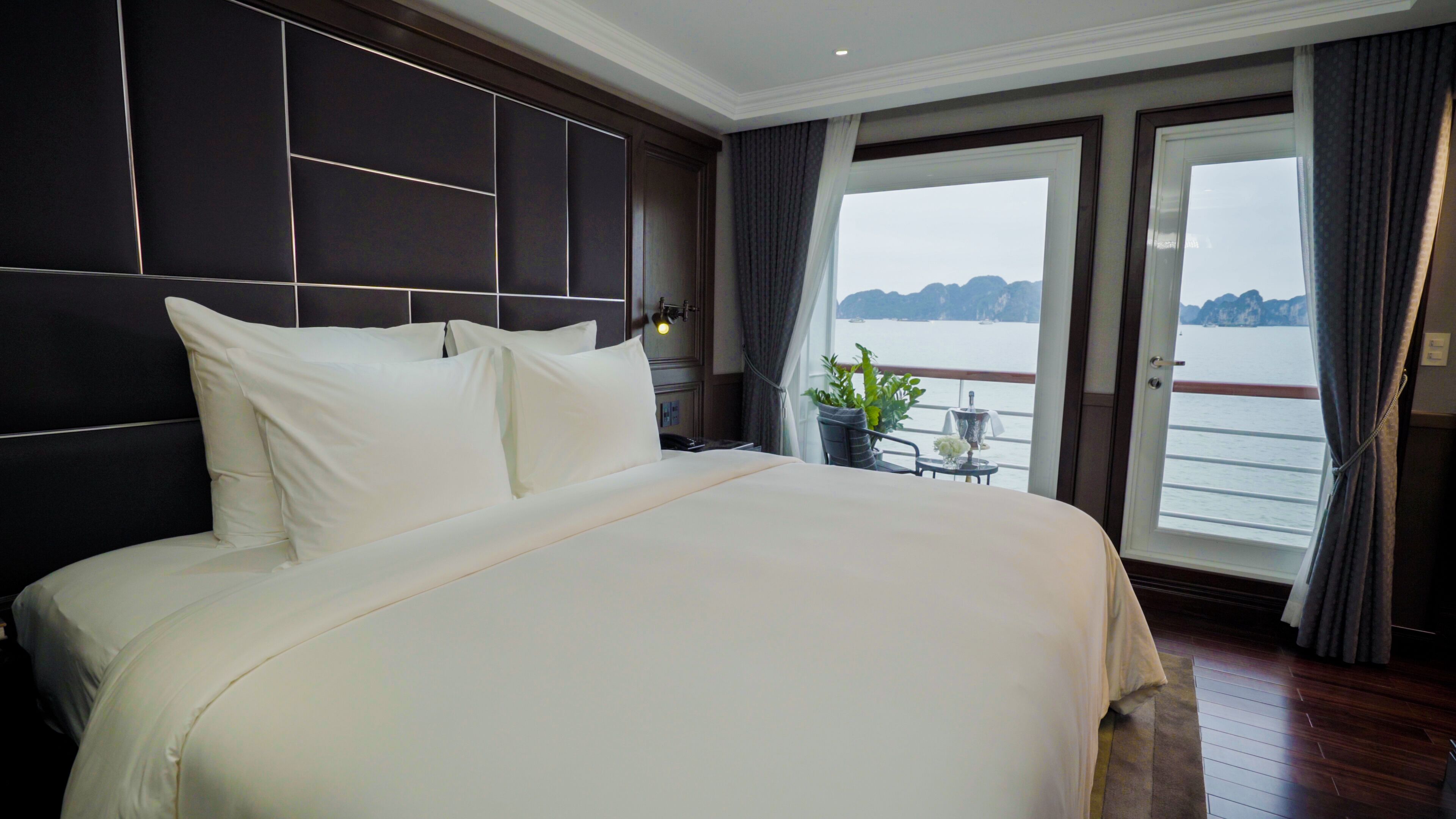 Ein großes Bett mit weißen Leinen in einem Zimmer, das ein dunkles Kopfteil, einen kleinen Tisch mit einer Pflanze und einen Blick auf das Wasser durch ein großes Fenster bietet.