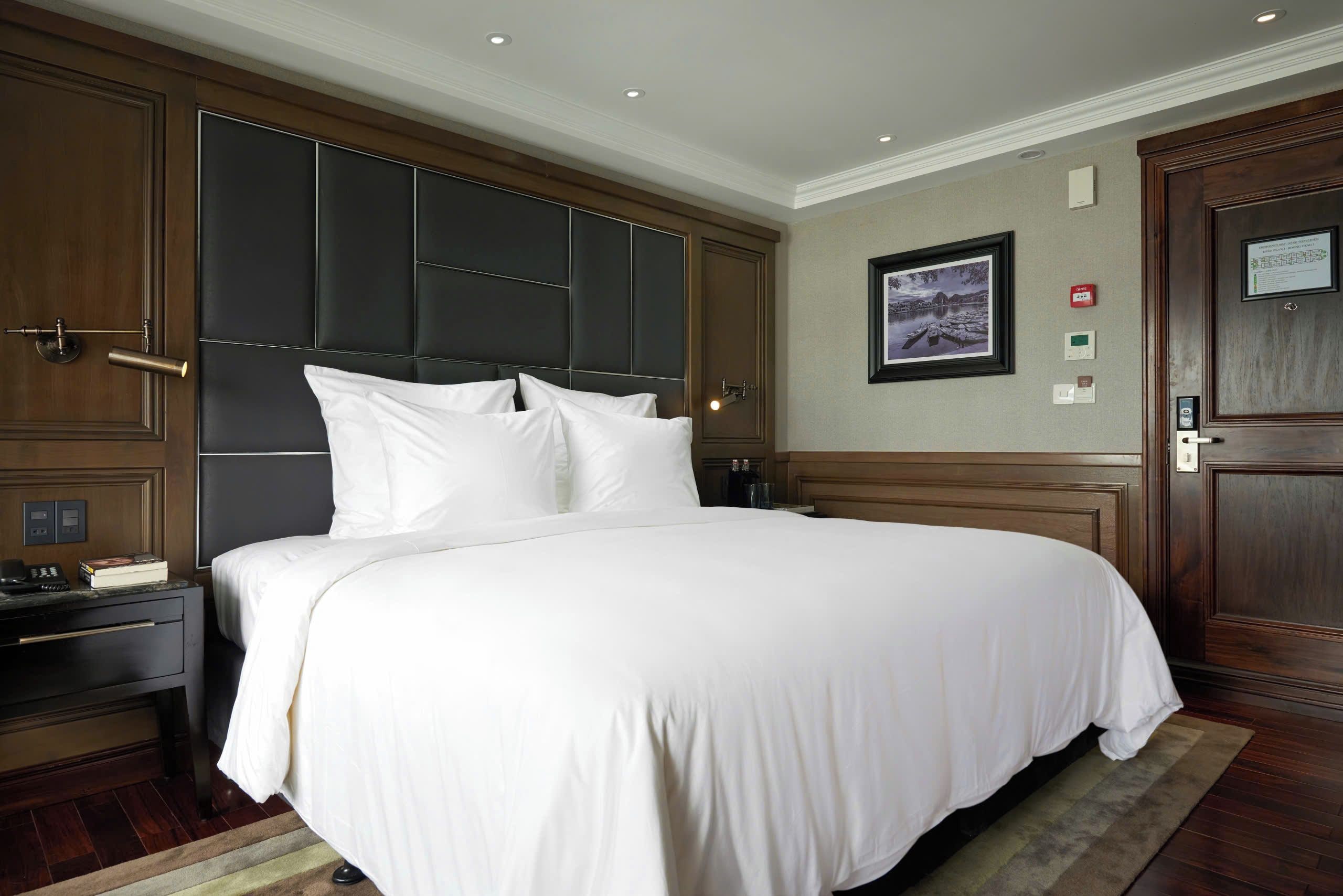 Une chambre d’hôtel avec un grand lit, une table de chevet avec un téléphone et des livres, un tableau encadré au mur et une porte dotée d’un panneau de sécurité.