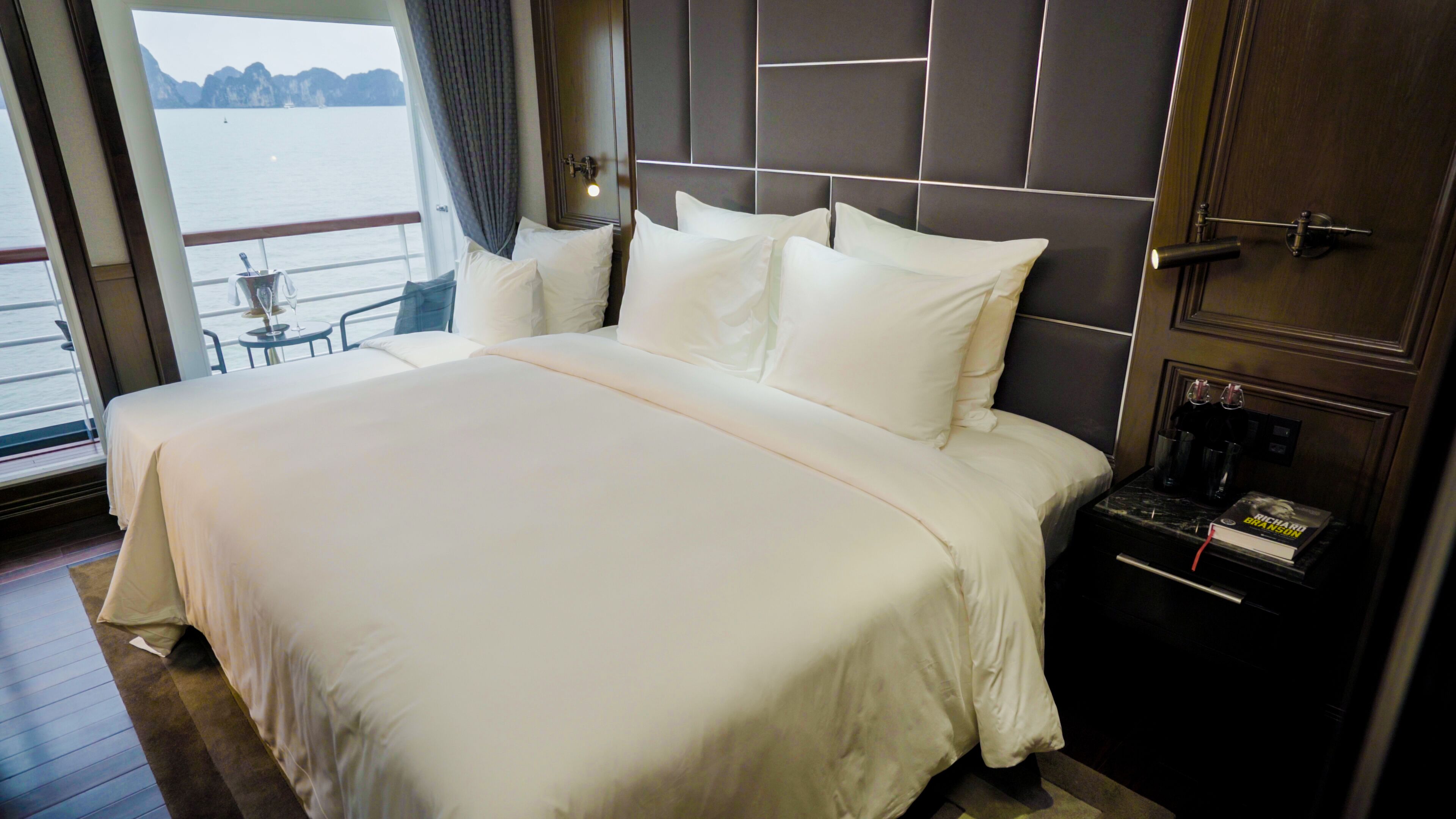Een groot bed met witte lakens, een nachtkastje met een boek, en uitzicht op het water door een raam.