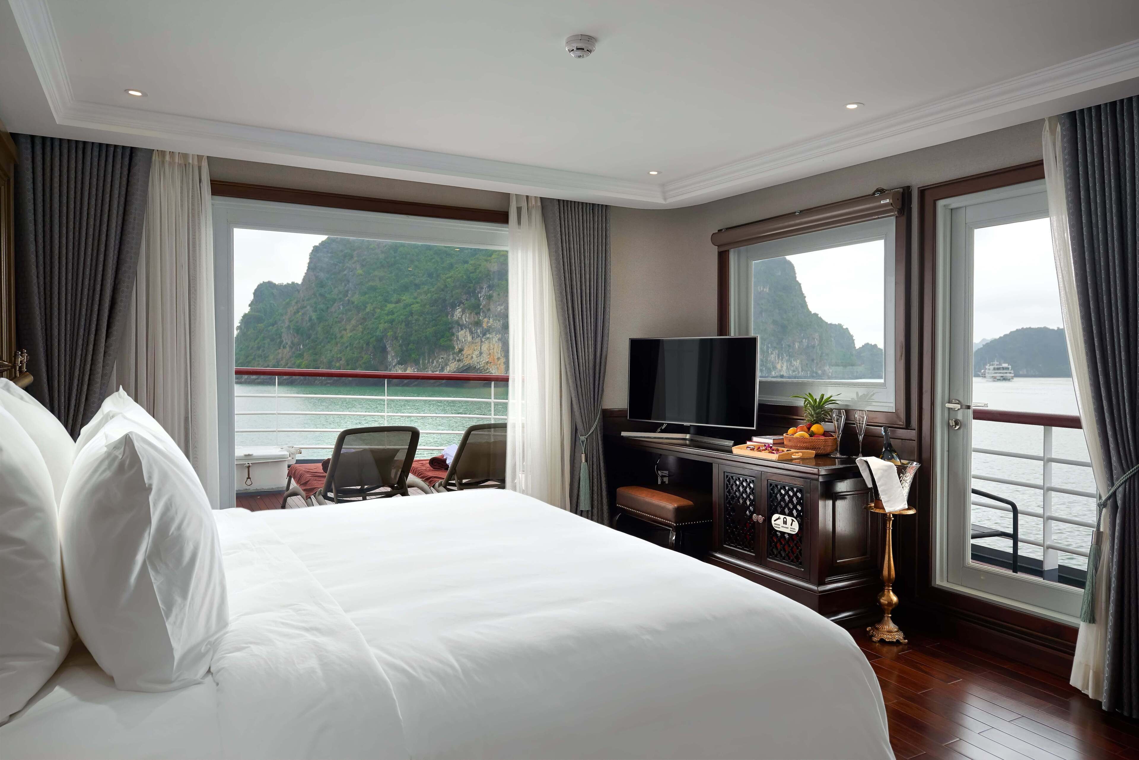 A hotel room with a large bed, a TV, a small table with fruit and a bottle, and a view of the sea and mountains.