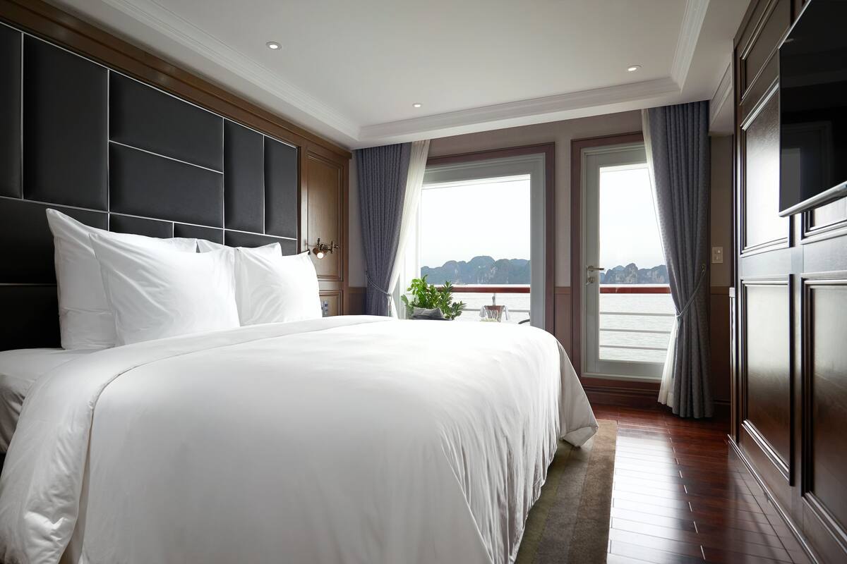 Ein großes Bett mit weißer Bettwäsche, ein dunkles Kopfteil und ein Blick ins Freie durch ein Fenster mit Vorhängen.