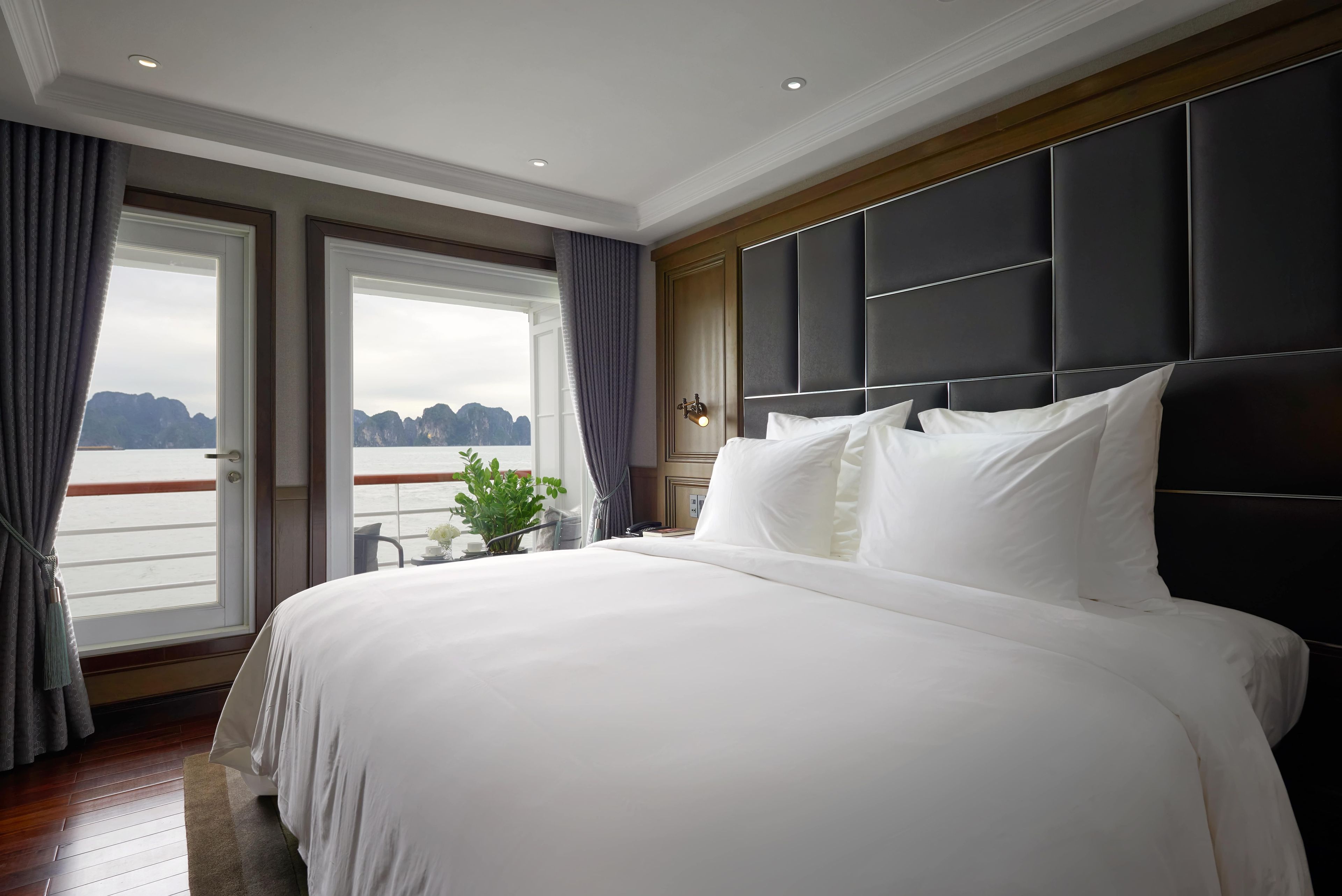 A large bed with a dark headboard, a view of the sea through a large window, and a small plant on the windowsill.