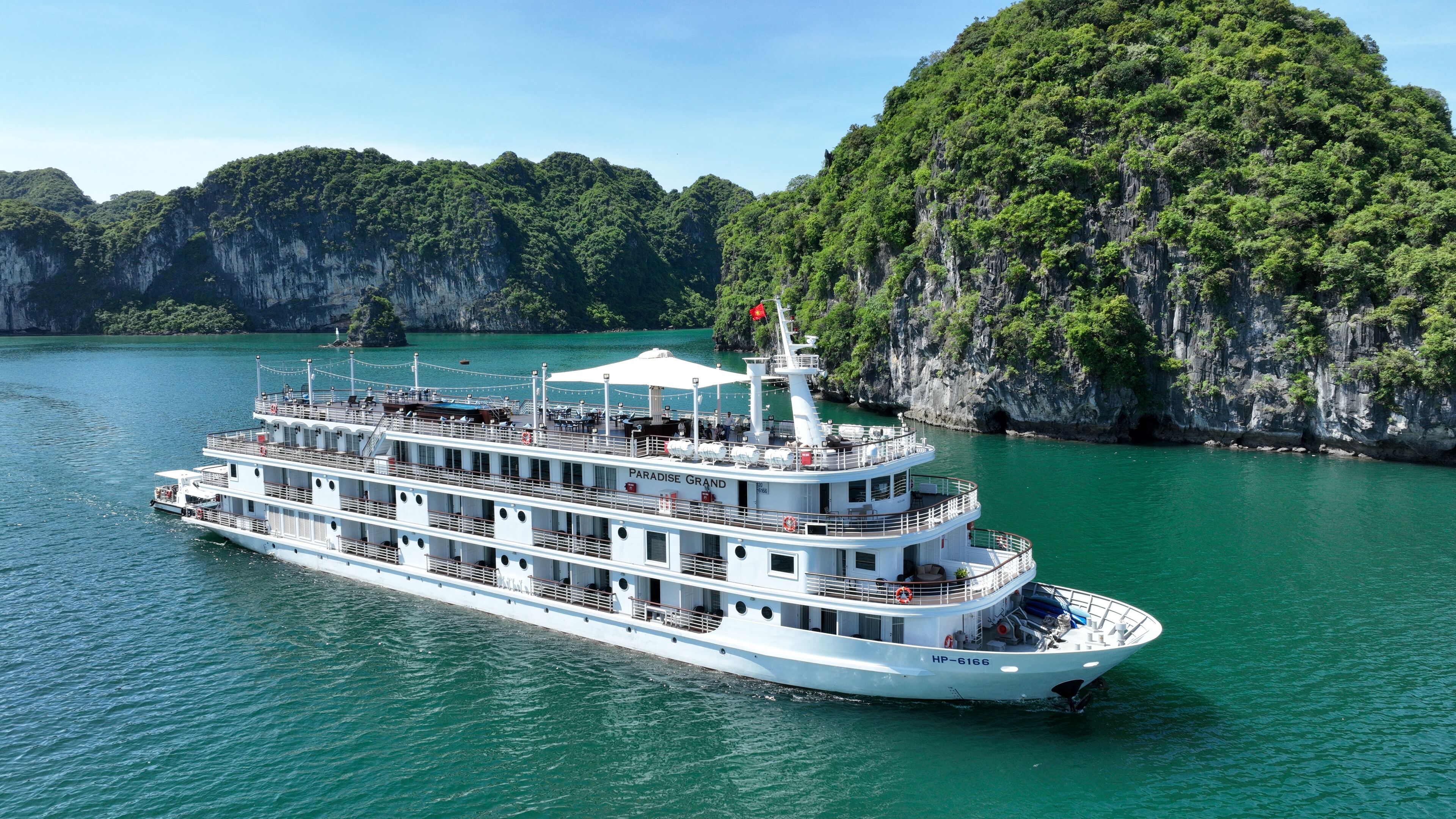 A large, multi-deck cruise ship with a white exterior and multiple levels of balconies, sailing in a turquoise sea with lush green cliffs in the background.