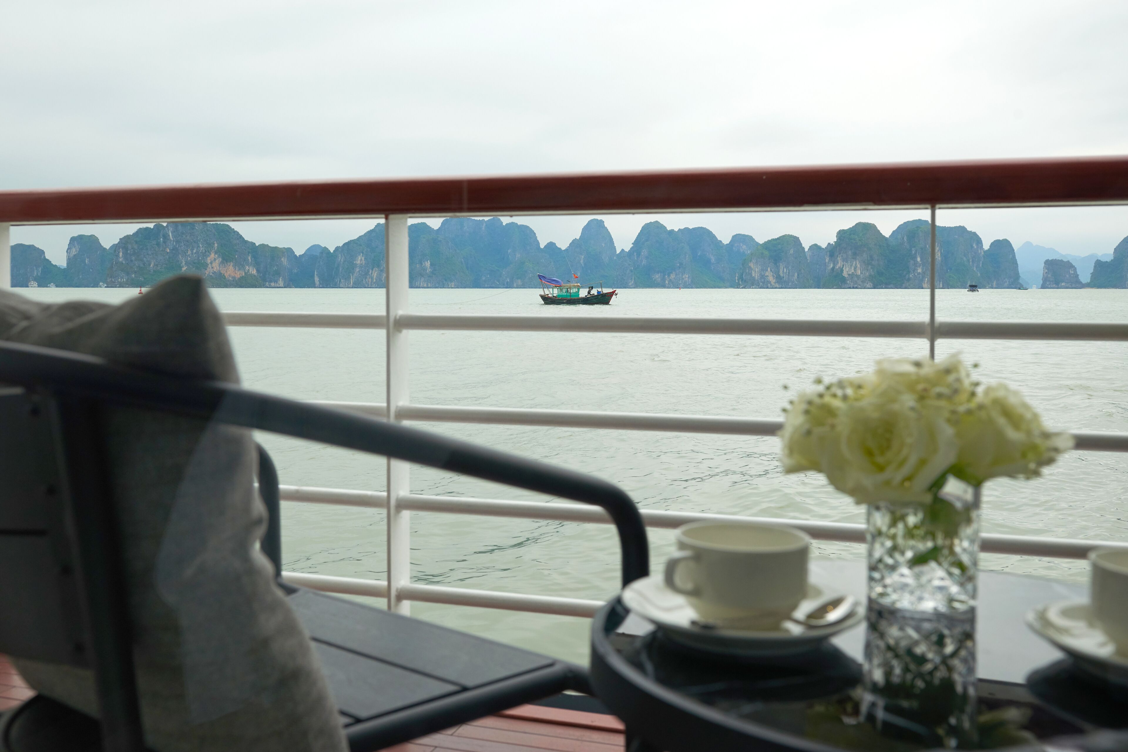 A table with a vase of white flowers and two cups, a chair, and a view of a boat on the water with distant rocky formations.