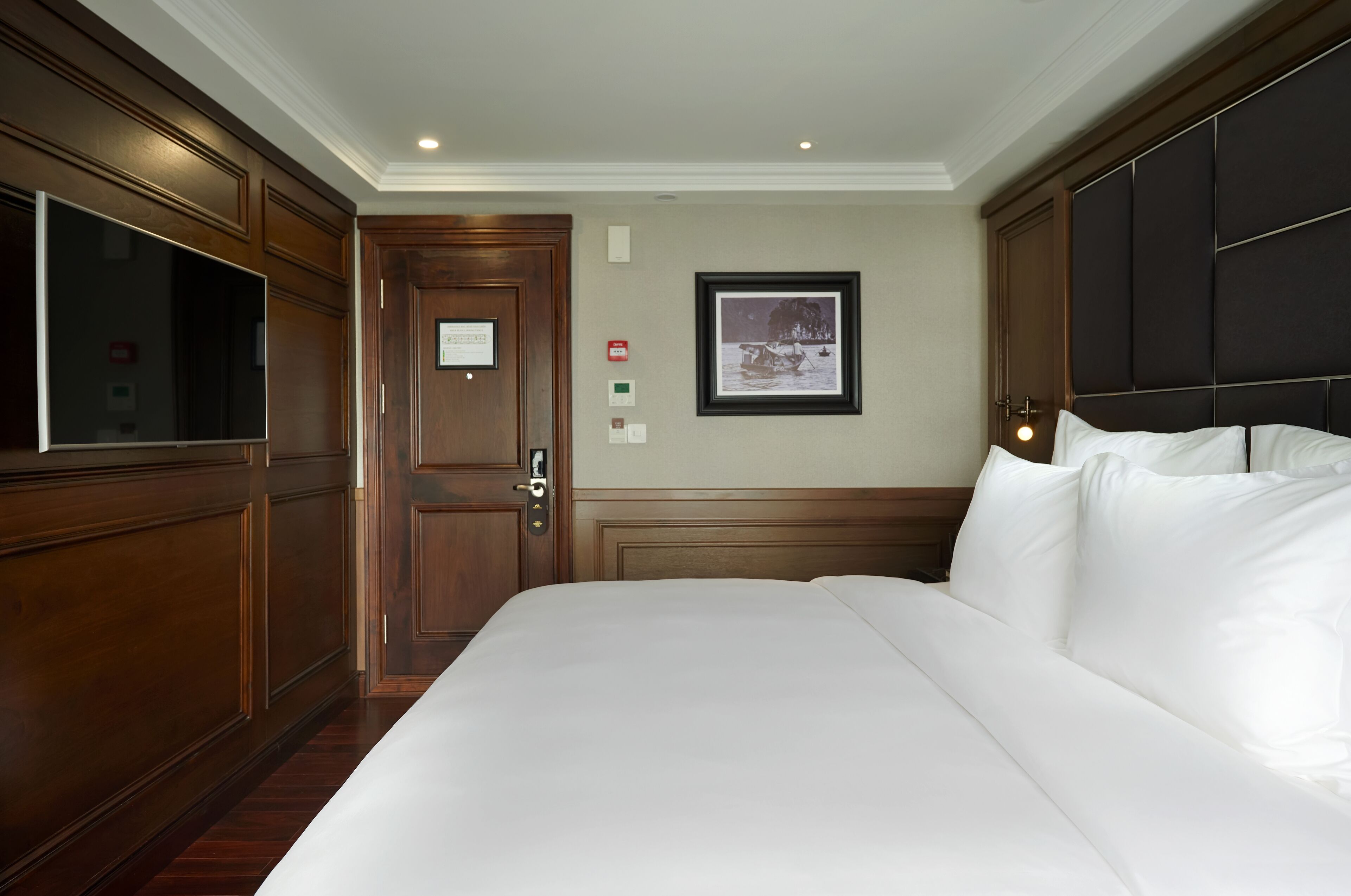 A hotel room with a large bed, wooden paneling, a flat-screen TV, a framed picture on the wall, and a door with a sign.