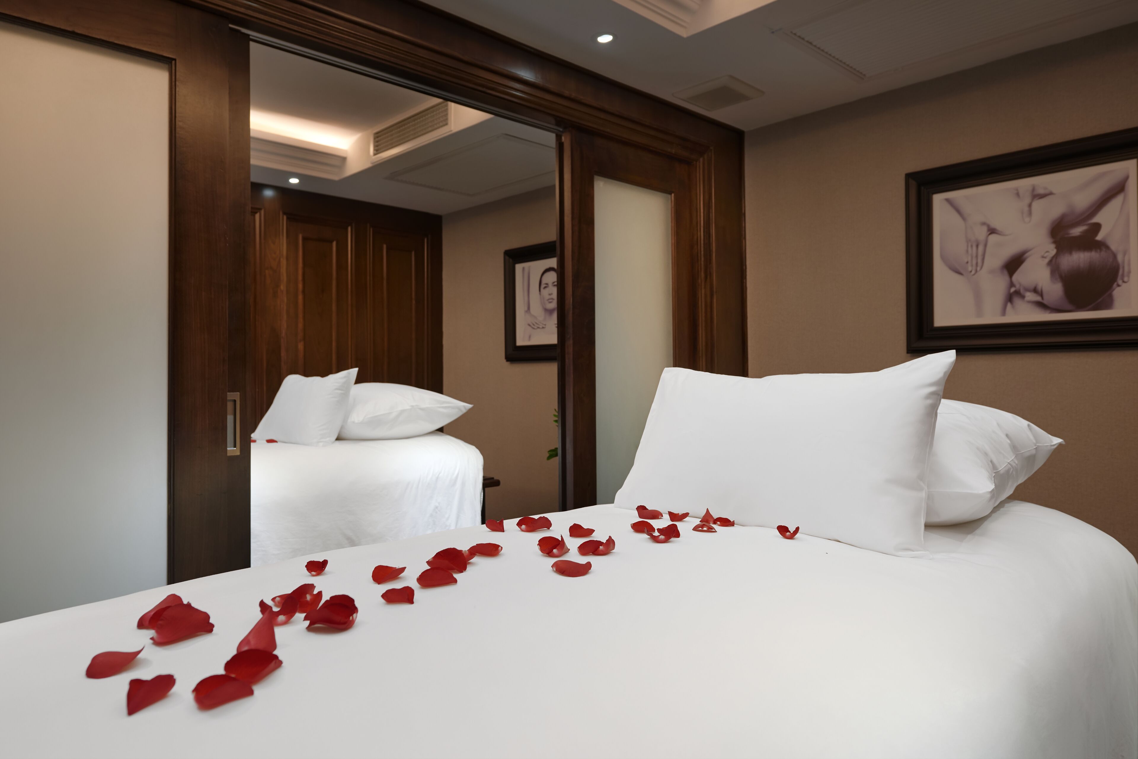 A hotel room with a large bed, white bedding, and red rose petals on the bed.