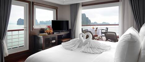 A hotel room with a large bed, a TV, a desk with a computer, and a view of the sea.