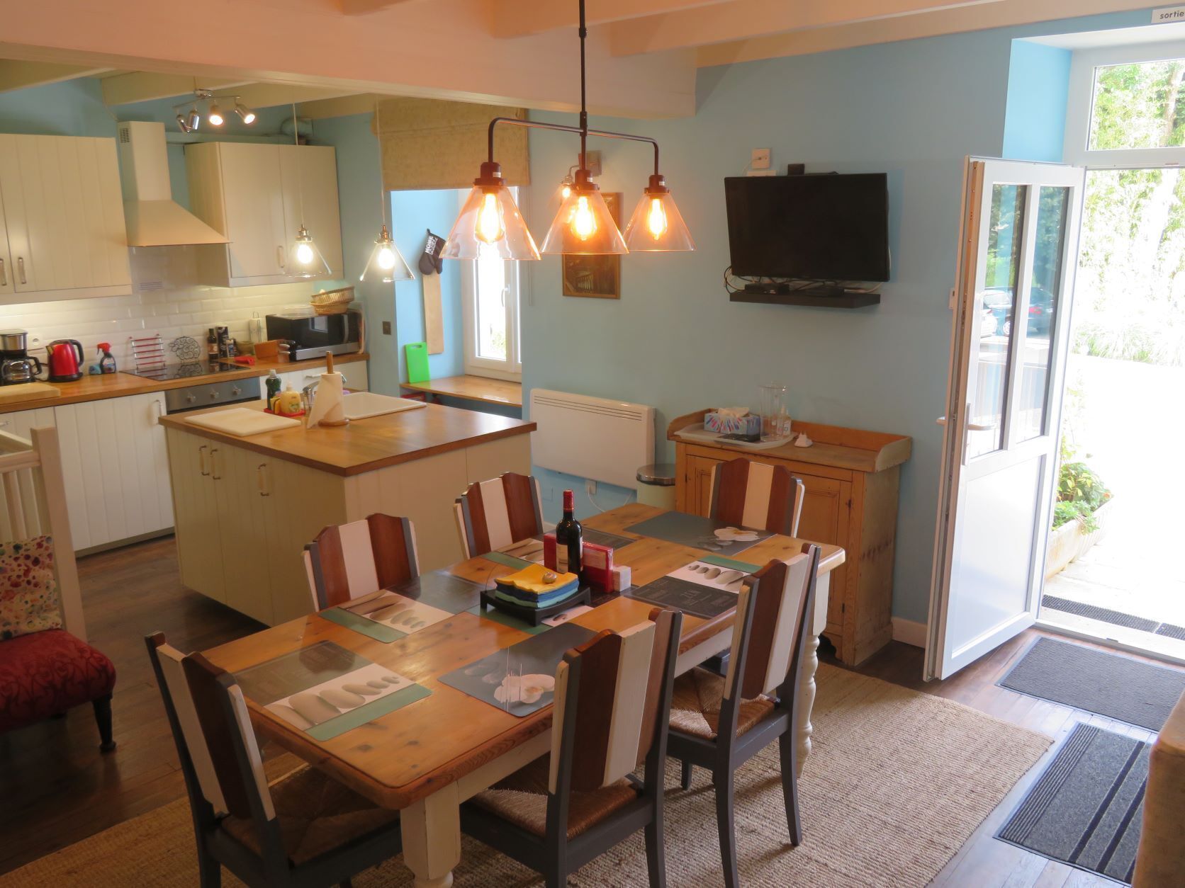 A modern kitchen with a central island, a dining area with a wooden table and chairs, and a TV mounted on the wall.