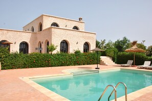 Zona de piscina con un edificio de piedra, cielo despejado y un jardín bien cuidado.