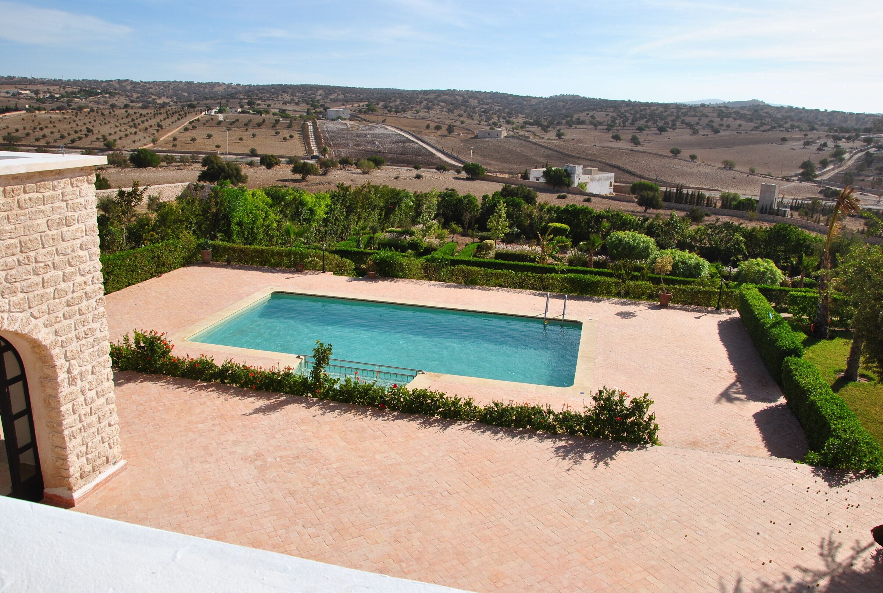 A swimming pool with a paved deck, surrounded by neatly trimmed hedges and a view of a rural landscape.