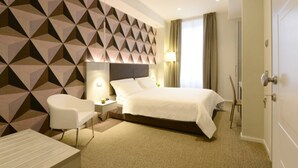 Une chambre d’hôtel moderne avec un mur orné d’un motif géométrique, un grand lit avec du linge de lit blanc, une table de chevet avec une lampe, un fauteuil blanc et une porte sur la droite.