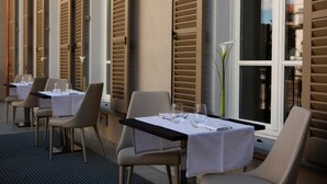 Un espacio de comedor al aire libre con mesas dispuestas para dos, con un diseño moderno, sillas y mesas de colores claros.
