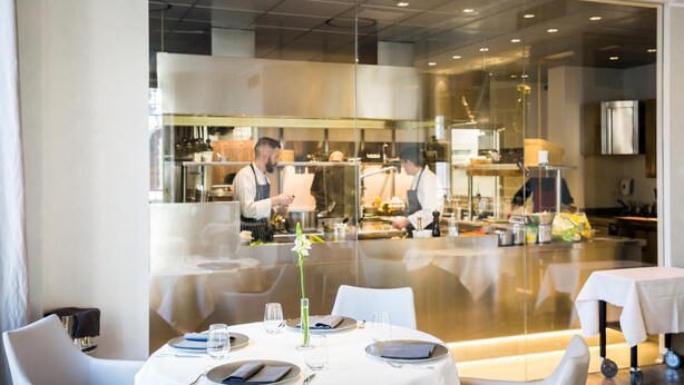 Un restaurant avec des tables dressées pour le service, une cuisine à l’arrière-plan et un décor intérieur moderne.