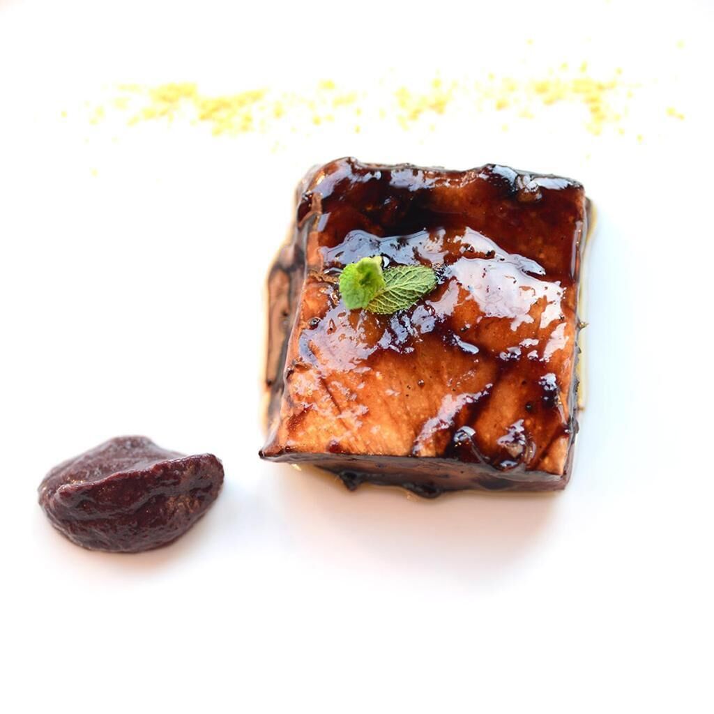 Un postre cuadrado con glaseado brillante y adorno de hoja de menta, acompañado de una única fruta oscura sobre una superficie blanca.