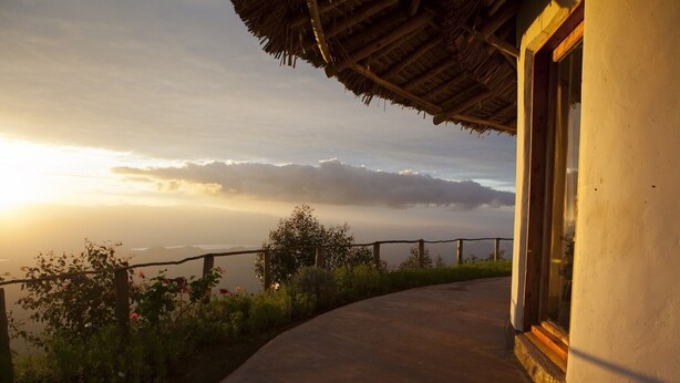 A curved pathway leading to a building with a thatched roof, overlooking a mountainous landscape with a sunrise or sunset.
