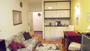 Un espace de vie compact comprenant un coin cuisine, un canapé avec des coussins, une table à manger recouverte d’une nappe rouge et un téléviseur.
