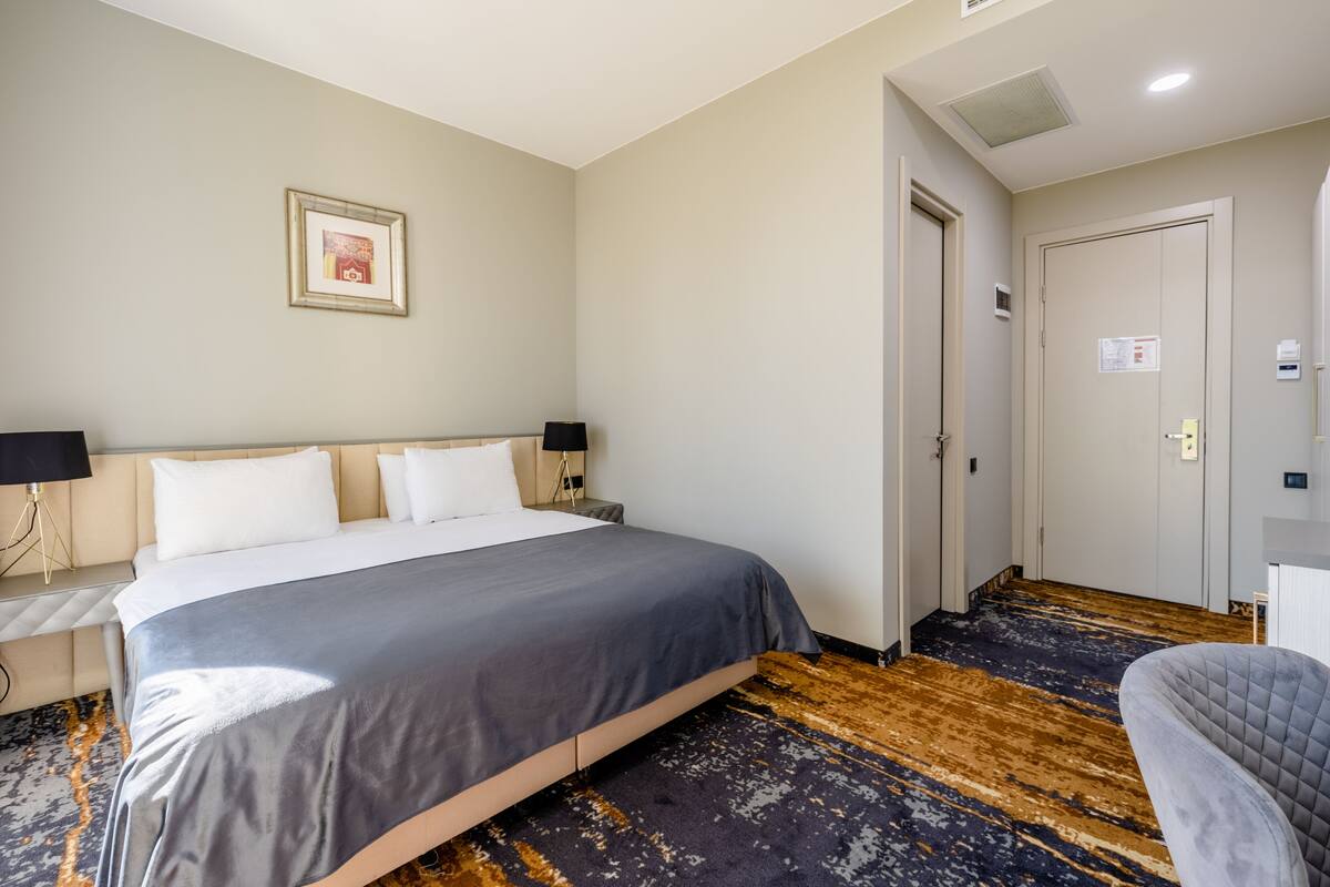 A hotel room with a large bed, a bedside table with a lamp, a framed picture on the wall, and a door leading to another room.