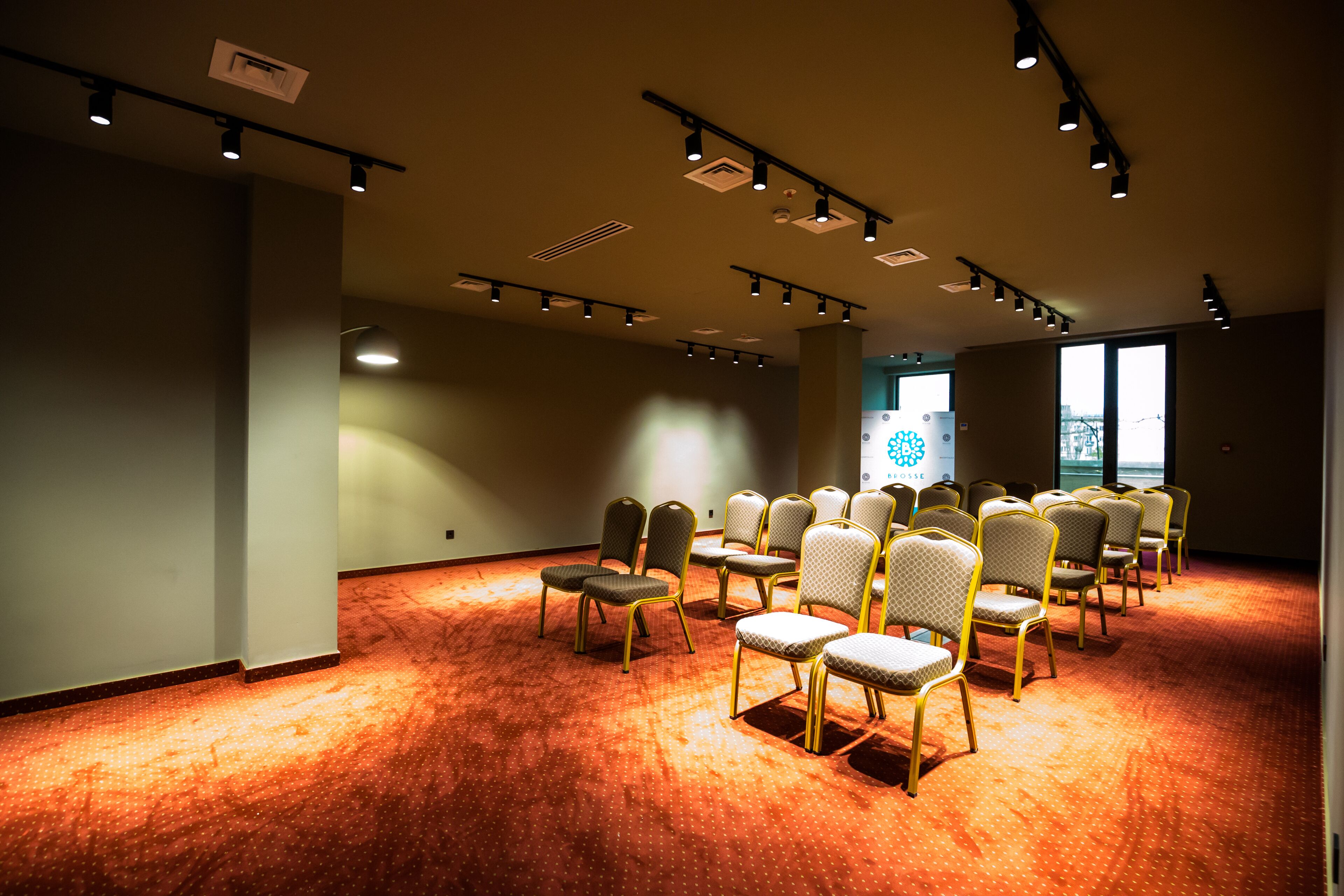 Un salón de conferencias con filas de sillas, luces en el techo y un ventanal grande.