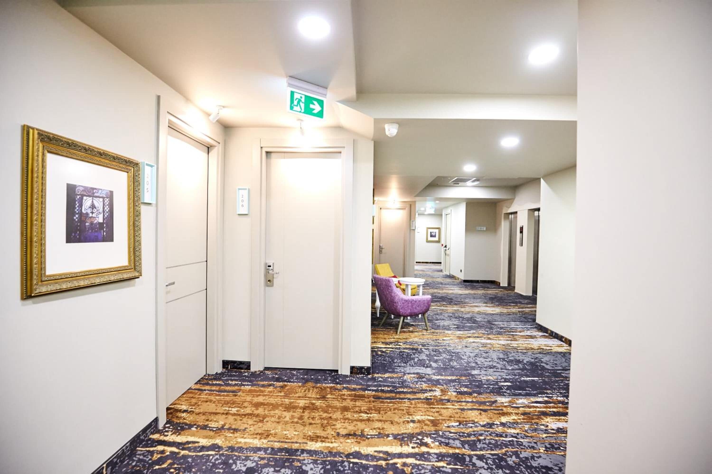 A hotel hallway with a patterned carpet, framed artwork on the walls, and a seating area with a purple chair and table.
