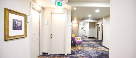 A hotel hallway with a patterned carpet, framed artwork on the walls, and a seating area with a purple chair and table.