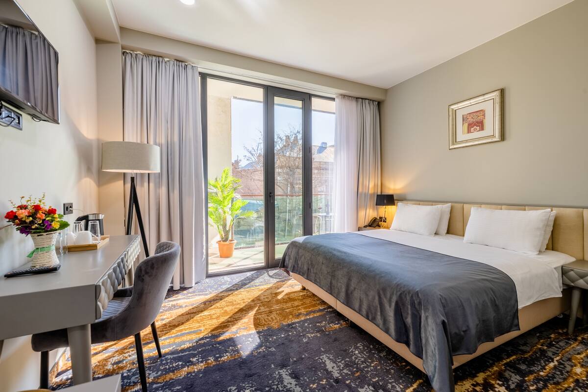 A hotel room with a bed, a desk with a lamp, a chair, a dining table with a vase of flowers, and a sliding glass door leading to a balcony with a plant.